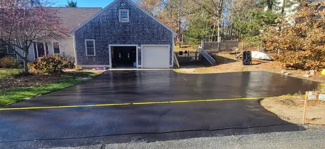 Freshly sealed black asphalt driveway in front of a house with autumn foliage in the background.